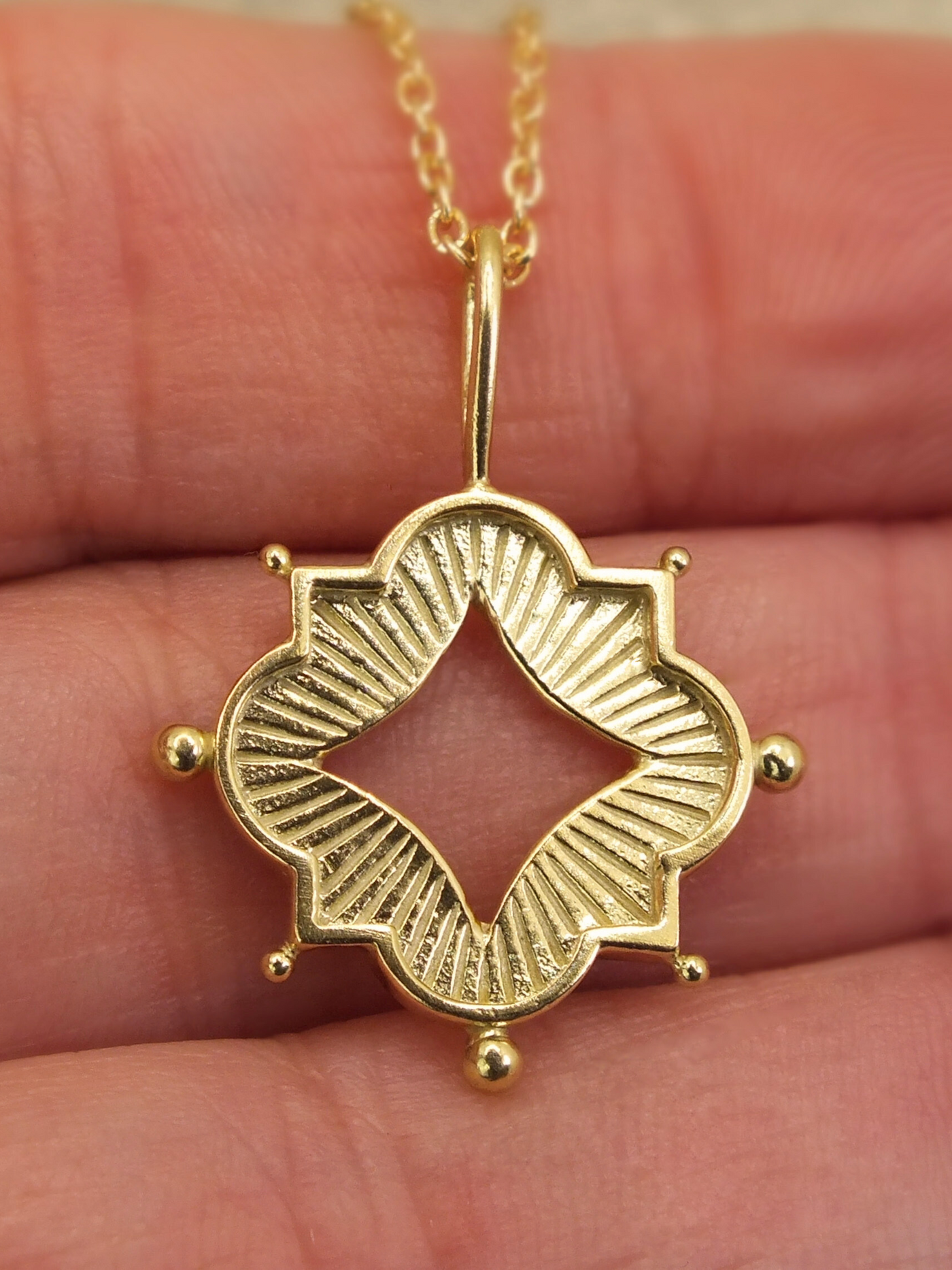 Gold quatrefoil pendant necklace with a central diamond-shaped cutout, inspired by Gothic architecture and sacred geometry, featuring engraved light rays and protective talisman-style detailing with spherical accents, hanging from a delicate gold chain