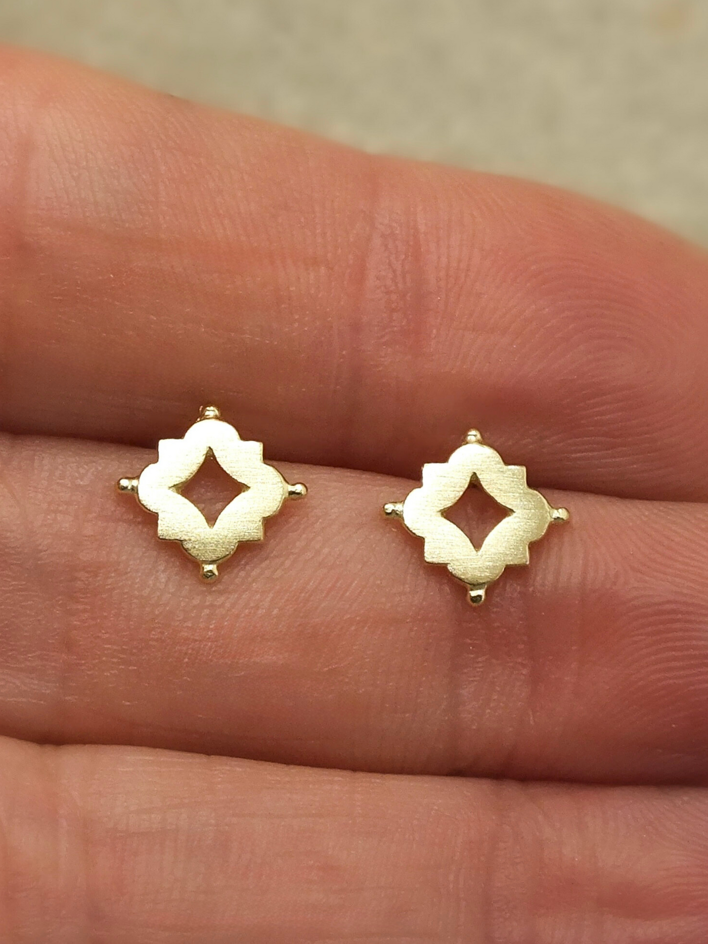 Pair of solid 18k gold stud earrings featuring a quatrefoil-inspired design with a central diamond-shaped cutout, accented by small granule-like details at each corner. The openwork creates a delicate window of light at the center, echoing the intricate tracery of Medieval and Renaissance architecture. A blend of historical elegance and minimalist form, perfect for everyday wear or timeless adornment