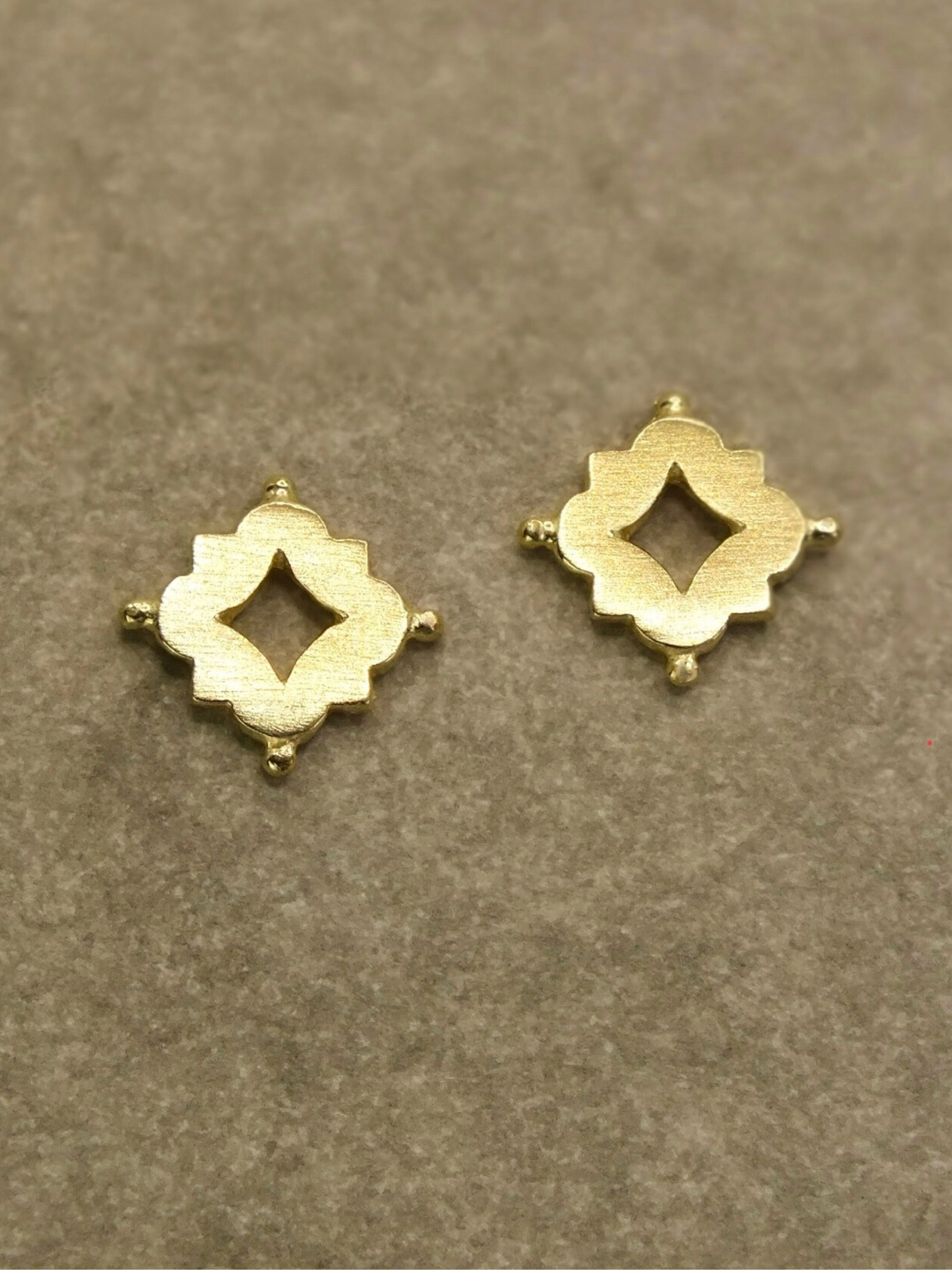Pair of small solid 18k gold stud earrings featuring a quatrefoil-inspired design with a central cutout, accented by small granule-like details at each corner. The motif draws from Medieval and Renaissance architectural ornamentation, blending historical elegance with minimalist form